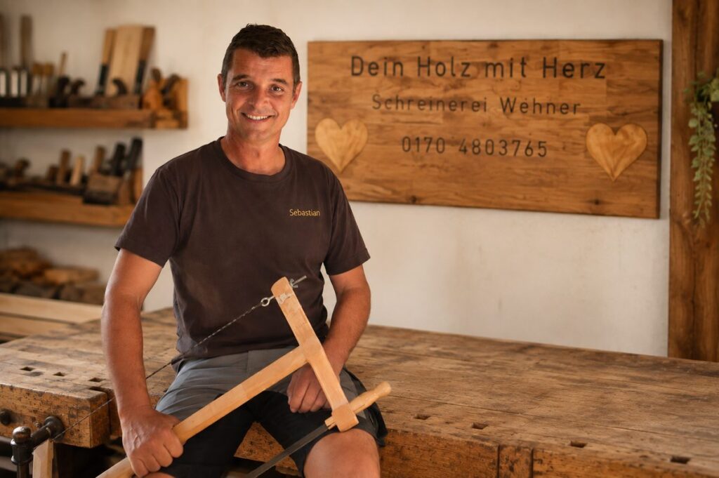 Schreiner Sebastian Wehner mit einer Säge in der hab, der in die Kamera lächelt. Im Hintergrund ein selbst erstelltes Holzschild mit dem Schriftzug "Dein Holz mit Herz" und der Telefonnummer 0170-4803765
